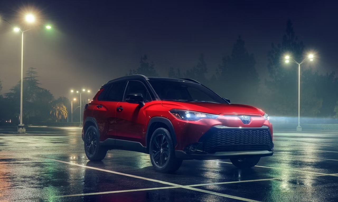 Vue trois quarts avant du Toyota Corolla Cross 2025 rouge stationné la nuit sous des lampadaires, mettant en avant son style robuste.