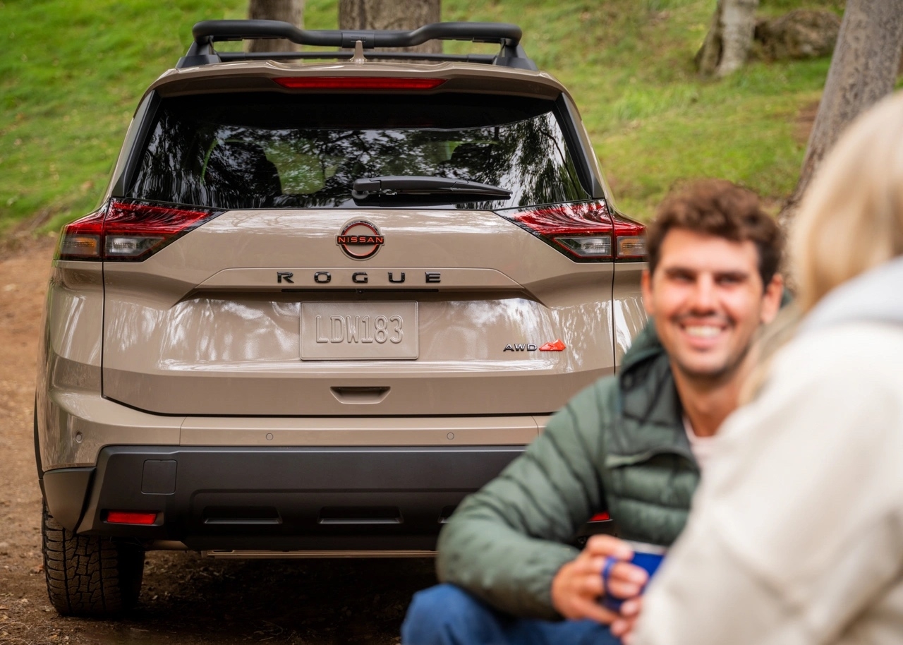 Vue arrière du Nissan Rogue 2025 beige en forêt avec un couple assis à proximité, illustrant son côté aventurier et convivial pour les escapades en plein air.